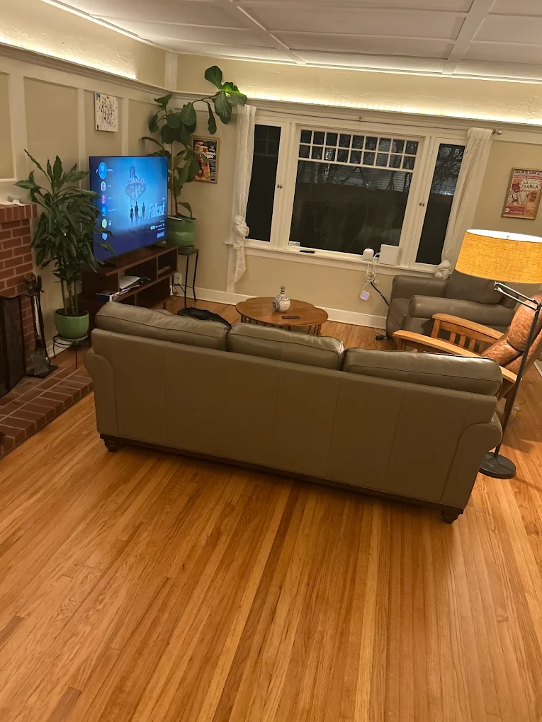 Warm oak strip flooring in a finished living room, seen from behind a gray leather sofa toward a brick fireplace.